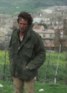 A man stands on a field, a city on the background.